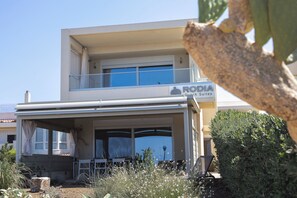 A modern two-story building with a flat roof and a balcony. The building has a sign that reads 'RODA Beach Suites.' There are outdoor chairs and a table visible in front of the building.