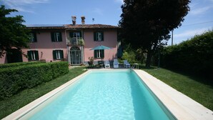 A rectangular swimming pool with a clear blue water surface, surrounded by a light-colored tiled deck. In the background, there is a pink building with green shutters and a balcony, and a green umbrella providing shade. - Eco-Villa with garden and swimming pool UNESCO Heritage area  (Govone)