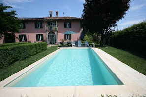 A rectangular swimming pool with a clear blue water surface, surrounded by a light-colored tiled deck. In the background, there is a pink building with green shutters and a balcony, and a green umbrella providing shade.