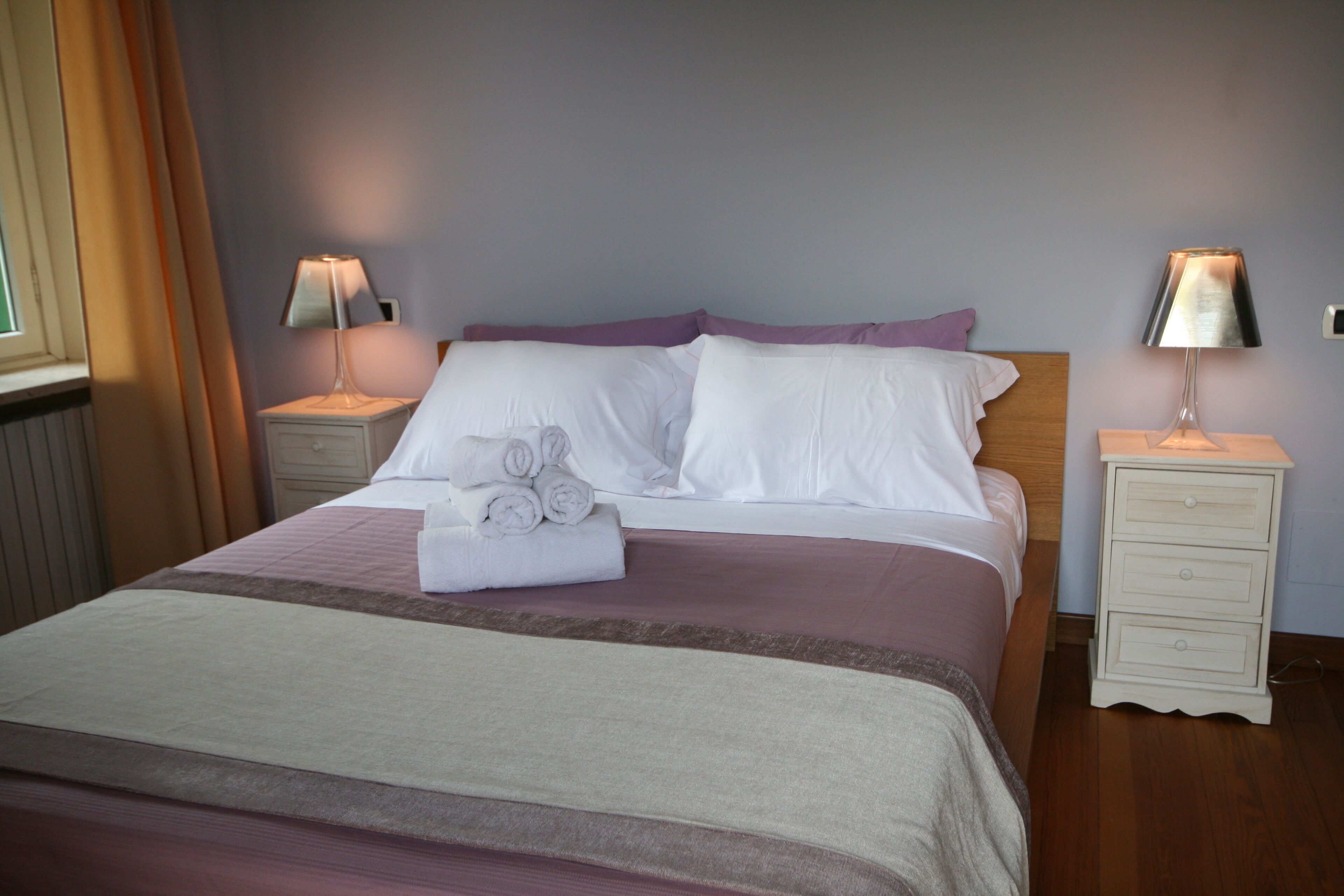 A neatly made bed with white linens, a purple pillow, and a set of rolled towels on the side. There are two bedside tables with lamps, and the room has a wooden floor.