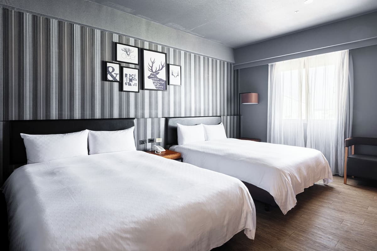 Habitación de hotel con dos camas, una mesita de noche con lámpara y un ventanal con cortinas.