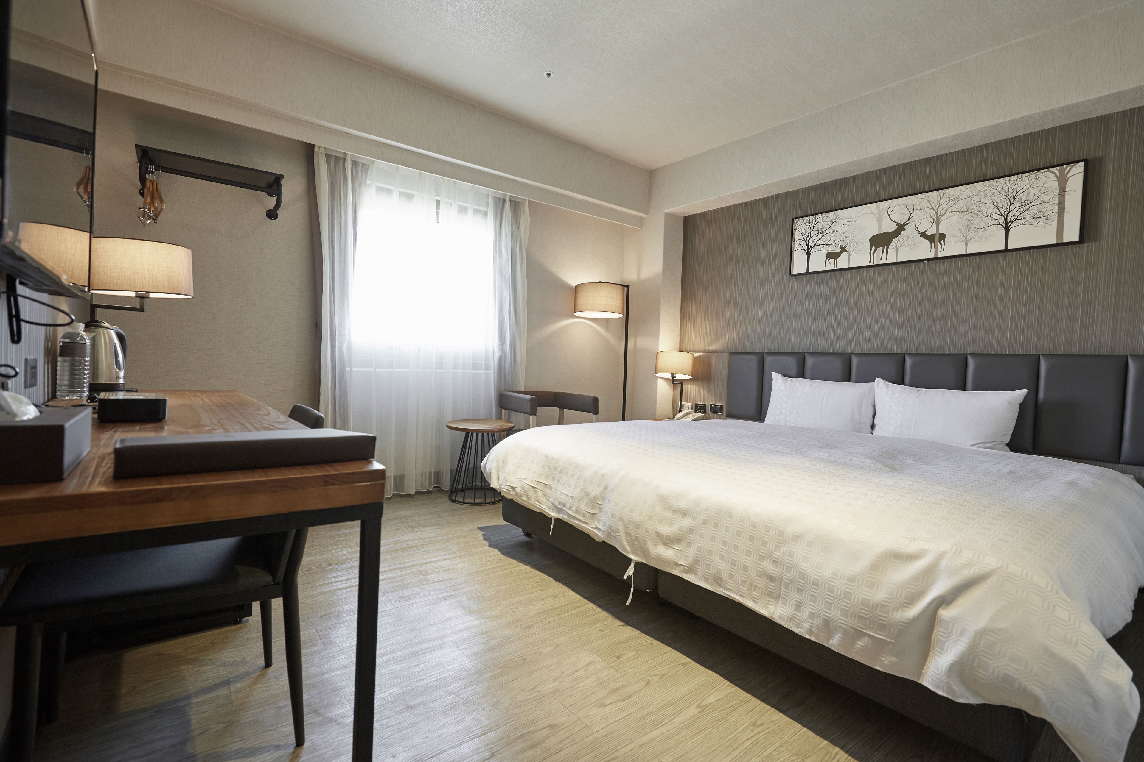 A hotel room with a large bed, a desk with a lamp, a small table, and a painting on the wall.