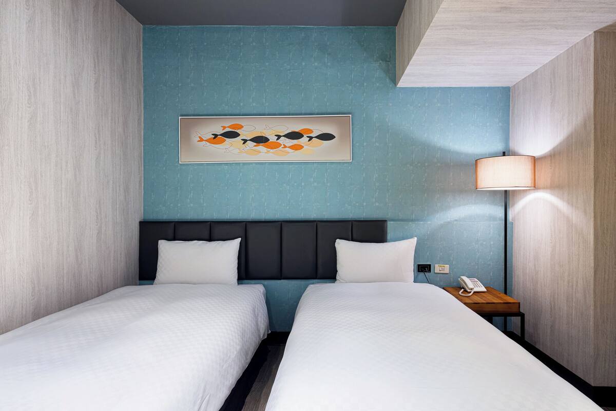 Habitación de hotel con dos camas, una pared azul decorativa, una obra de arte enmarcada, una mesita de noche con lámpara y un teléfono.