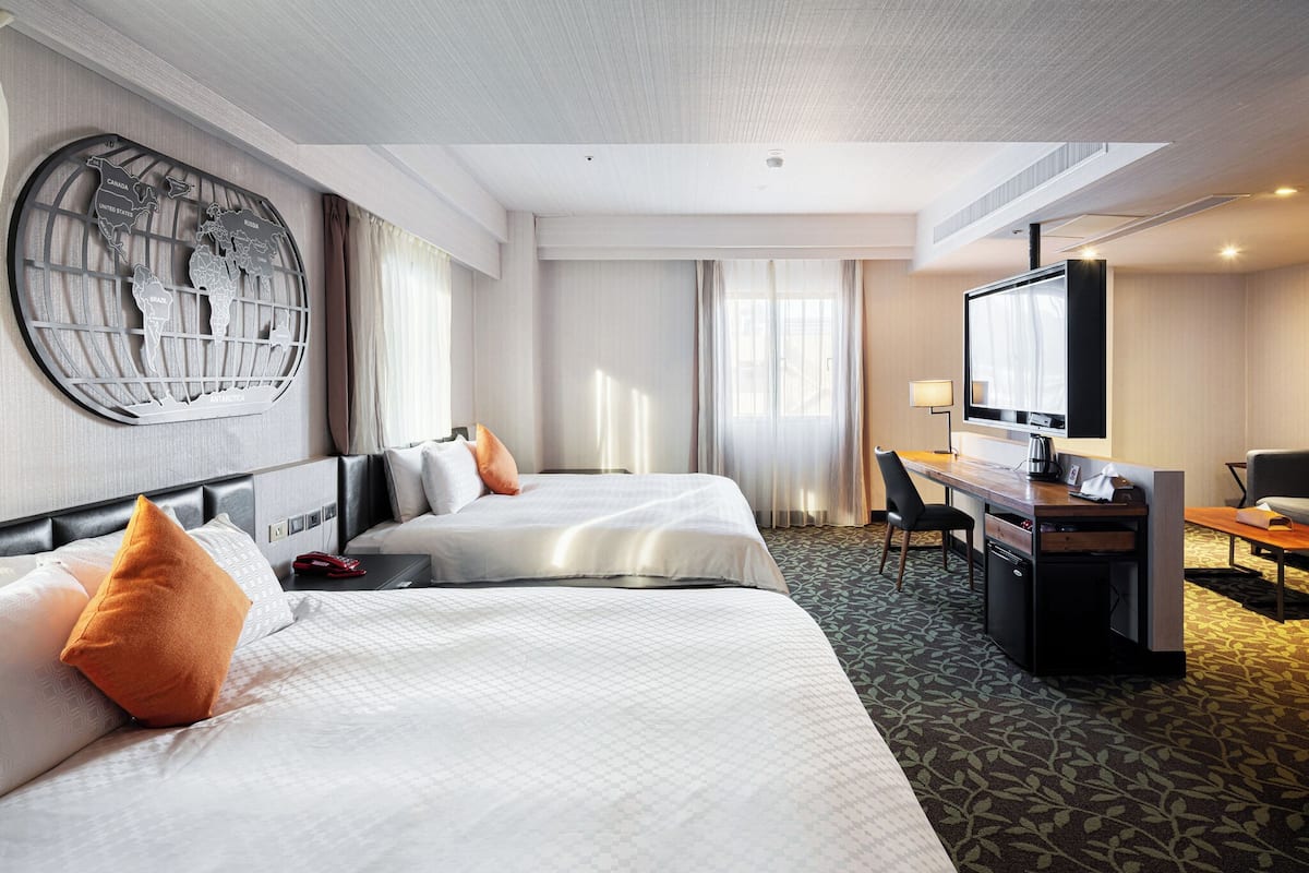 Habitación de hotel con dos camas, un escritorio, un televisor y un gran mapa mundial en la pared.