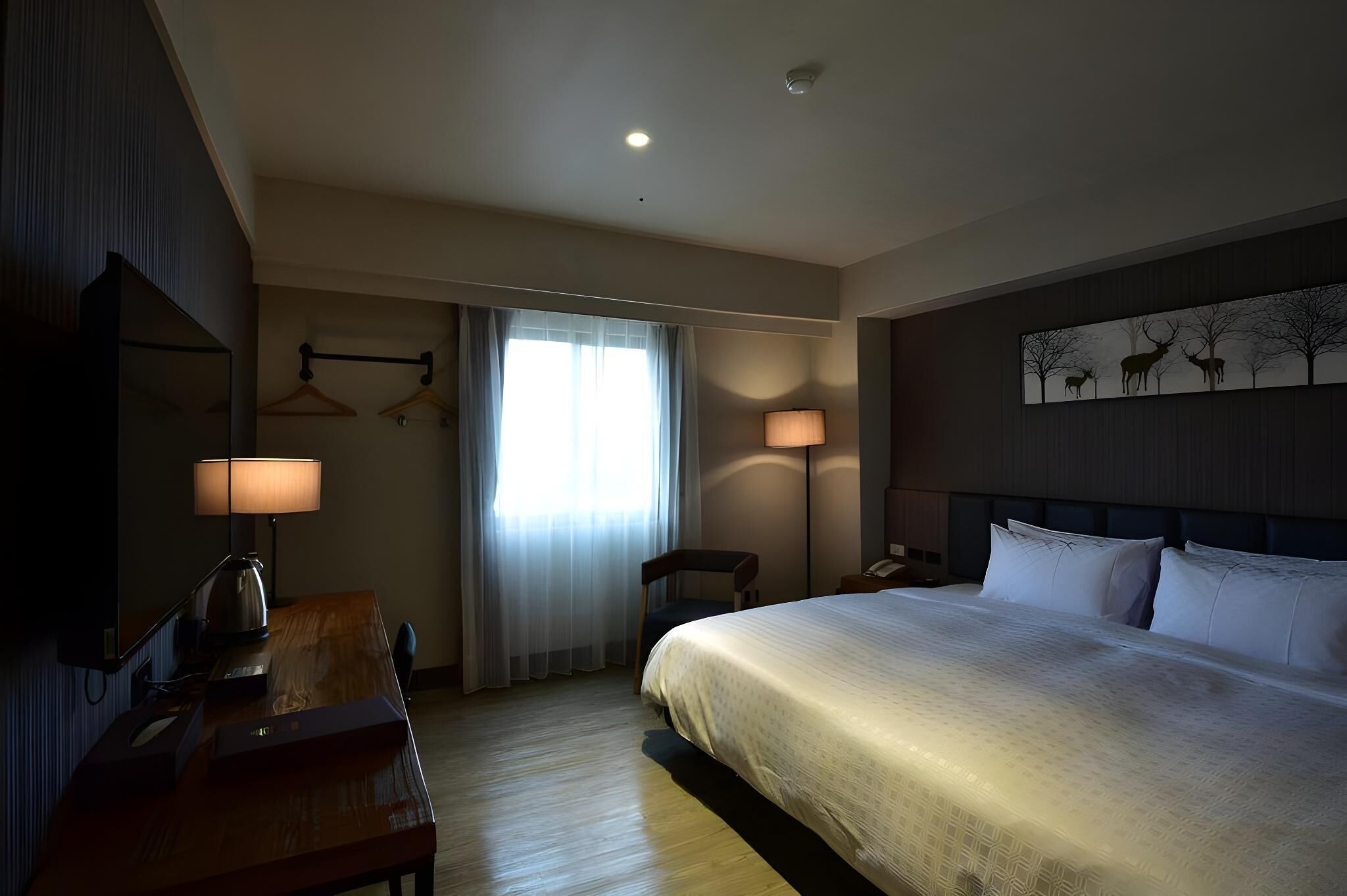 A hotel room with a large bed, a desk with a lamp, a chair, a desk lamp, and a wall-mounted artwork.