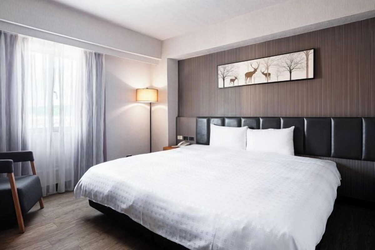 A modern hotel room with a large bed, a chair, a lamp, and a wall-mounted artwork.