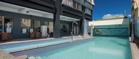 A swimming pool with a tiled deck, a covered seating area with chairs, and a building with large windows and a mural.