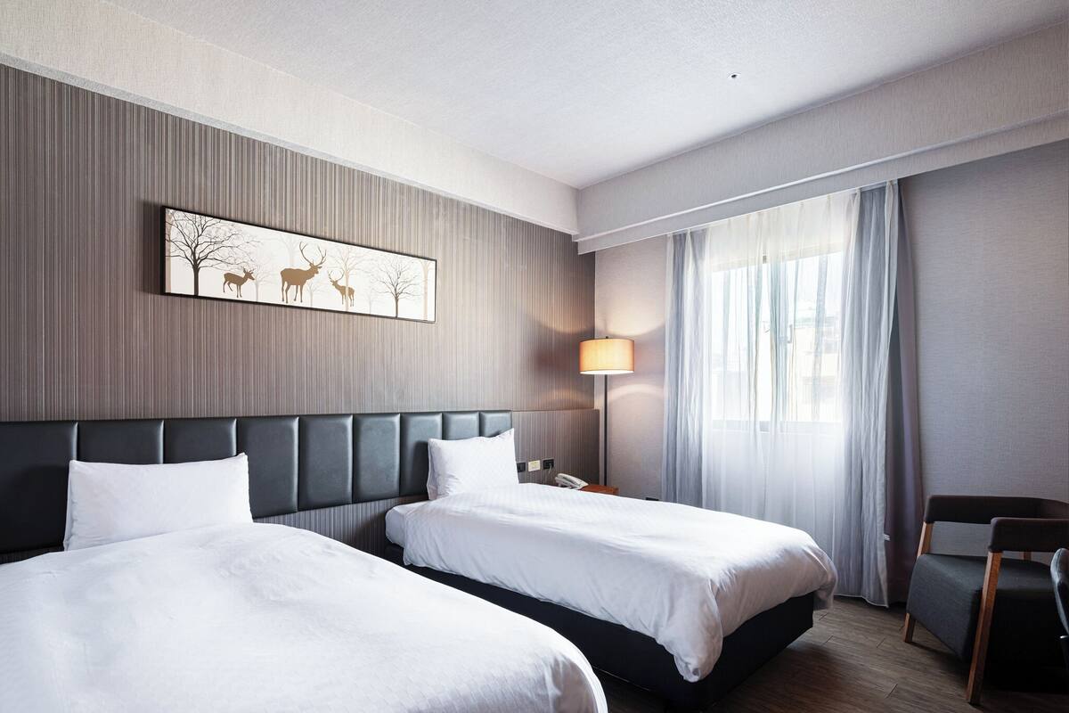 Habitación de hotel con dos camas, una mesita de noche, una lámpara, una silla y una ventana con cortinas.