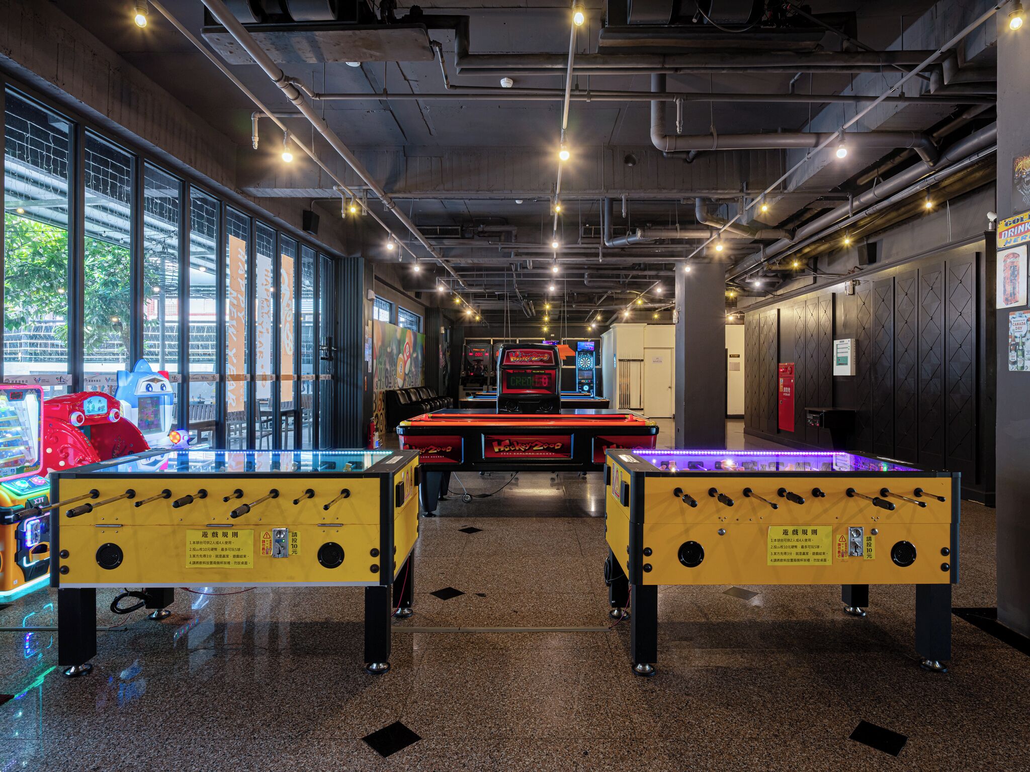 An indoor arcade with two foosball tables, arcade machines, and a large window with a view of the outside.