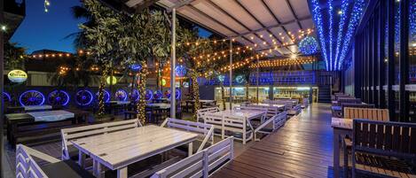 An outdoor dining area with wooden tables and benches, string lights, and a covered section with blue ambient lighting.