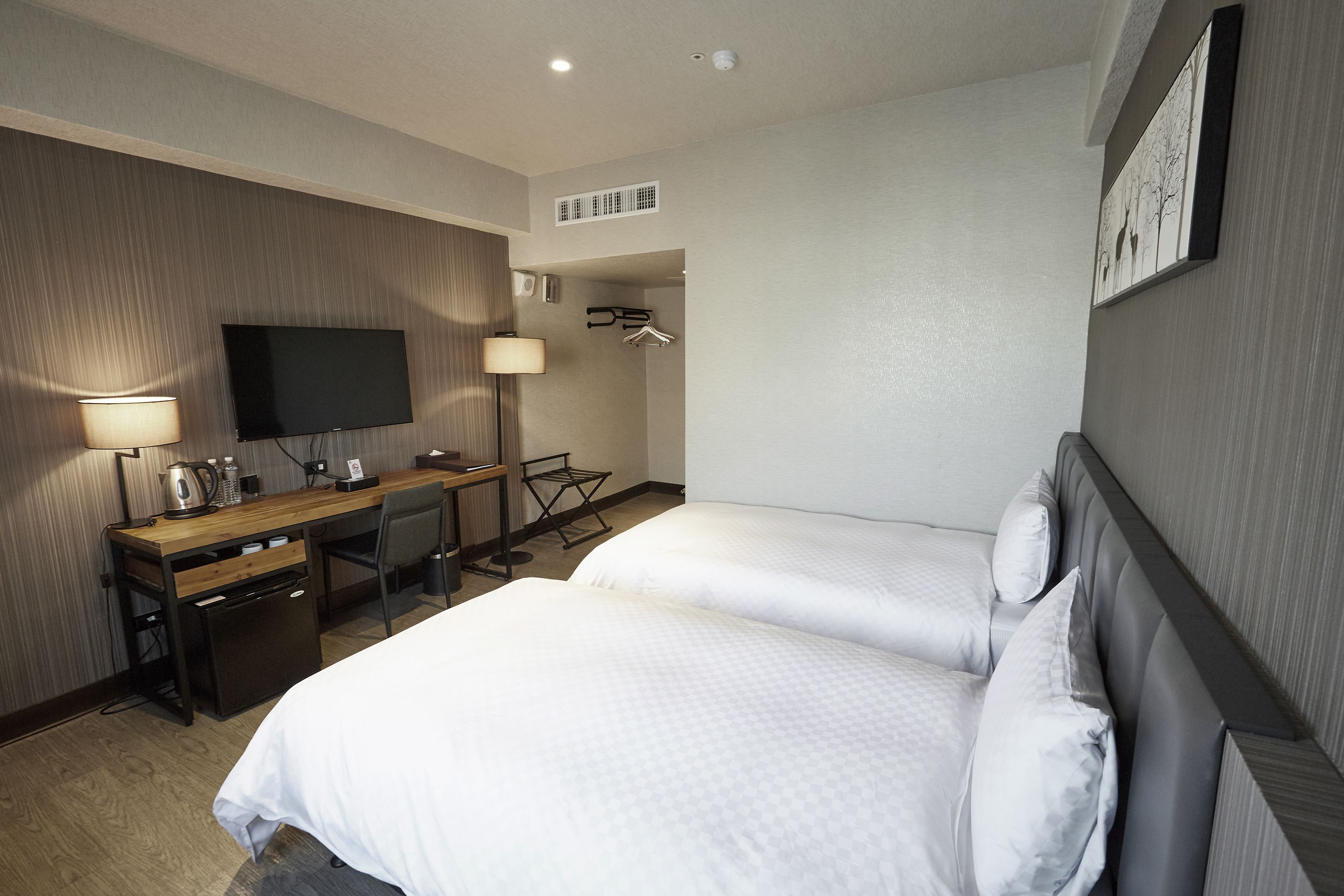 A hotel room with two beds, a desk with a flat-screen TV, a lamp, and a coat rack.