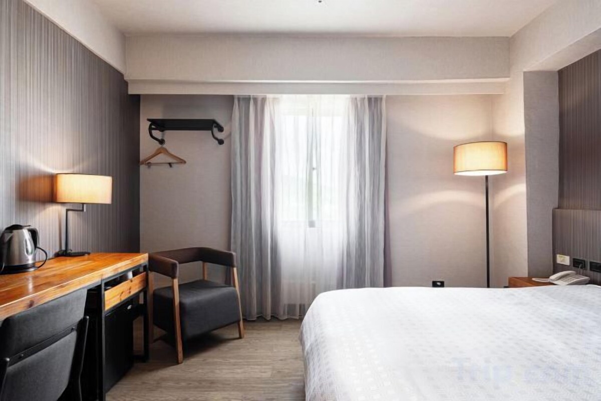 A hotel room with a bed, desk, chair, lamp, and a view of the outdoors.