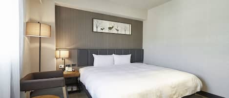 A hotel room with a bed, a desk with a lamp, a chair, and a small table.