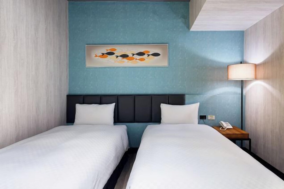 A hotel room with two beds, a dark headboard, a wall-mounted artwork, a bedside lamp, and a phone on a wooden nightstand.