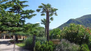 Une entrée fermée par un portail, avec un chemin de gravier menant à un bâtiment, le tout entouré d’une végétation luxuriante et dominé par une chaîne de montagnes en arrière-plan.