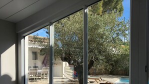A view through a large window of a backyard with a wooden deck, a tree, and a swimming pool.