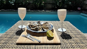 A wicker table with a plate of oysters, two glasses of champagne, a fork, a knife, and a half-cut lime by a pool.