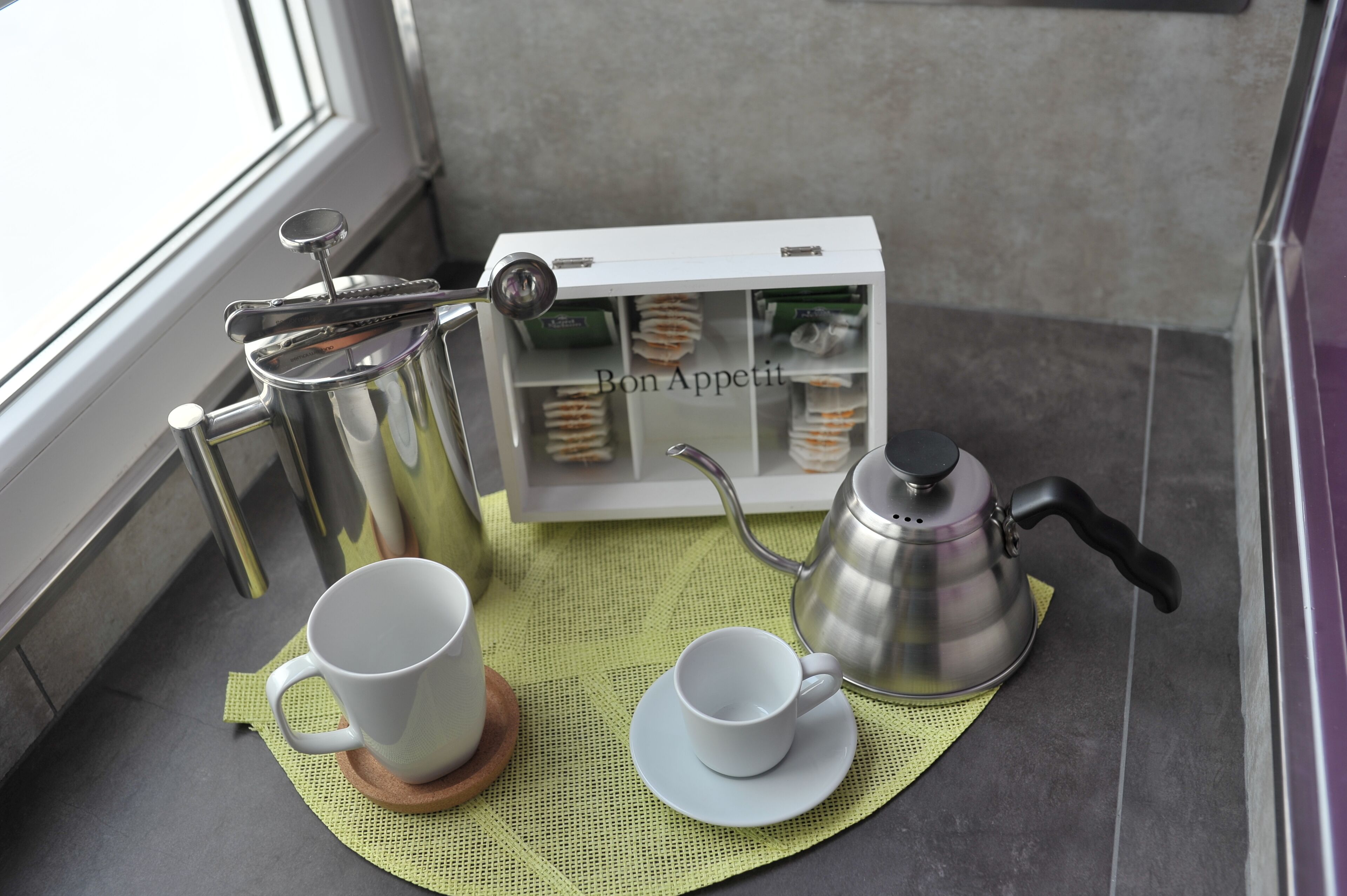 A tea setup with a teapot, cups, and a box labeled "Bon Appetit" on a tiled floor near a window.