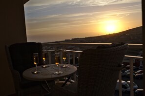 Un balcon avec une table dressée pour deux personnes, offrant une vue sur le paysage urbain au coucher du soleil.