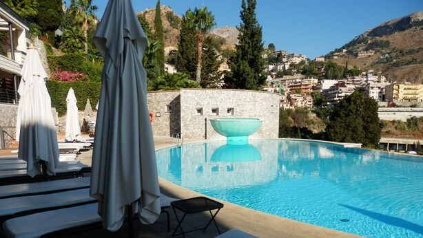 A swimming pool with lounge chairs and umbrellas, surrounded by stone walls and greenery, with a view of a hillside town in the background. - Fantastic apartment, Taormina with swimming pool, 5 minutes from Greek theater  (Taormina)