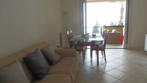 A living room with a beige sofa, a glass dining table, and chairs. There is a sliding glass door leading to an outdoor area with a red chair and a plant. - Fantastic apartment, Taormina with swimming pool, 5 minutes from Greek theater  (Taormina)