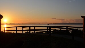Auringonlaskunäkymä puukannelta, jolta avautuu tyyni vesistö, ja kannen kaiteella istuu lintu.