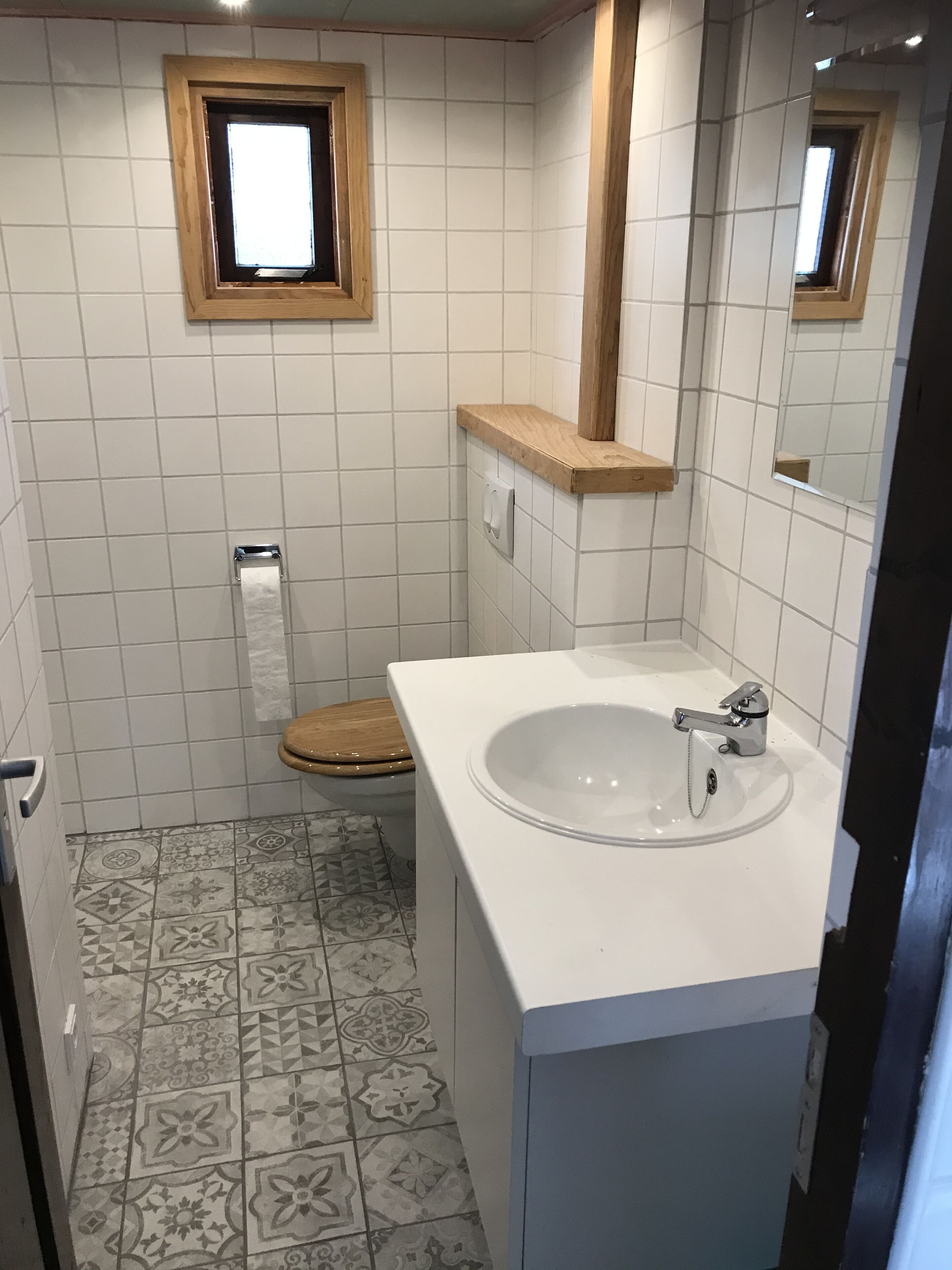 Un baño con un lavabo blanco, inodoro de madera y baldosas de suelo estampadas.