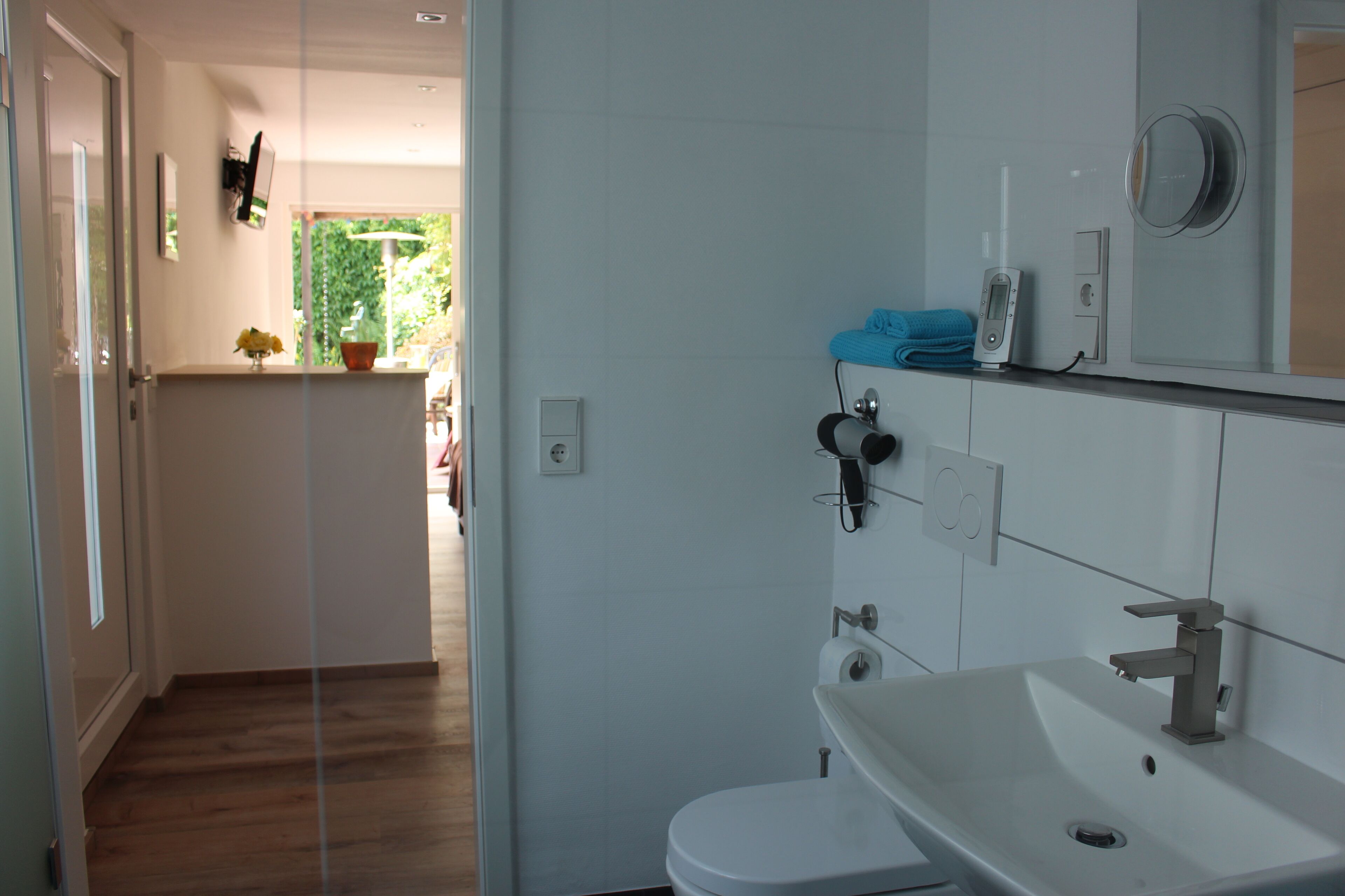 Un baño moderno con un lavamanos blanco, un inodoro y una zona de ducha. Hay un espejo sobre el lavamanos y un perchero con una toalla colgada.