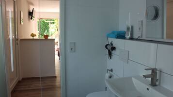 Un baño moderno con un lavamanos blanco, un inodoro y una zona de ducha. Hay un espejo sobre el lavamanos y un perchero con una toalla colgada.