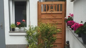 A potted plant in front of a wooden door, a window with a flower box, and a row of potted plants on the wall.