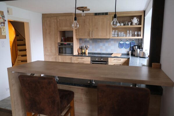 A modern kitchen with wooden cabinets, a central island with bar stools, hanging light fixtures, and a visible staircase.