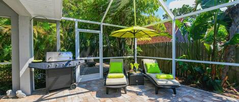 Un patio exterior con una parrilla, dos sillones de descanso con cojines, un parasol colgante y una piscina.