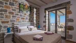 A bedroom with a stone wall, a bed with a patterned bedspread, a nightstand with a blue lamp, and a view of the sea through a glass door. - Charlise Luxury Villa by Cretevasion (Kokkini Hani)