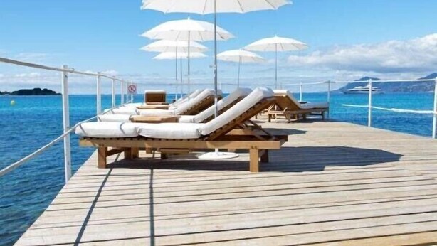 Una terrazza in legno con poltrone relax e ombrelloni bianchi, con vista su un mare calmo.