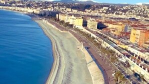 Una vista aerea di una città costiera con un’ampia spiaggia, un lungomare alberato da palme e edifici sullo sfondo.