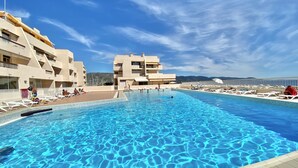 A swimming pool with clear blue water, surrounded by lounge chairs and a building with balconies.