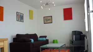 Een moderne woonkamer met een bruine bank, een glazen bijzettafeltje en een kleurrijk vloerkleed. Er hangen kunstwerken in lijsten aan de muren en een televisie op een dressoir.