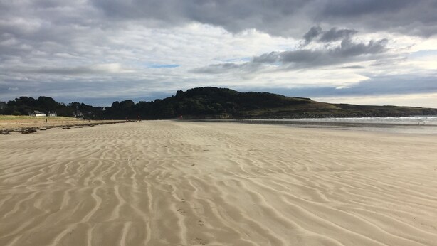 Een breed zandstrand met rustige golven, een verre heuvel en een bewolkte lucht.