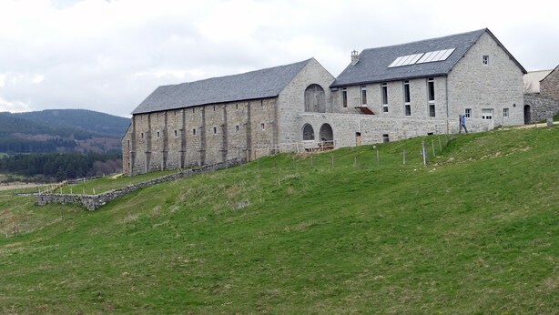 Un bâtiment en pierre, doté d’un toit en pente et de grandes fenêtres, situé sur une colline verdoyante et offrant une vue sur les collines lointaines.
