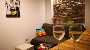 Un'accogliente zona living con un divano, un poggiapiedi e un tavolino in legno con due bicchieri di vino bianco. Un cuscino colorato a righe è appoggiato sul divano, e un'opera d'arte raffigurante una scena di strada in bianco e nero adorna la parete.
