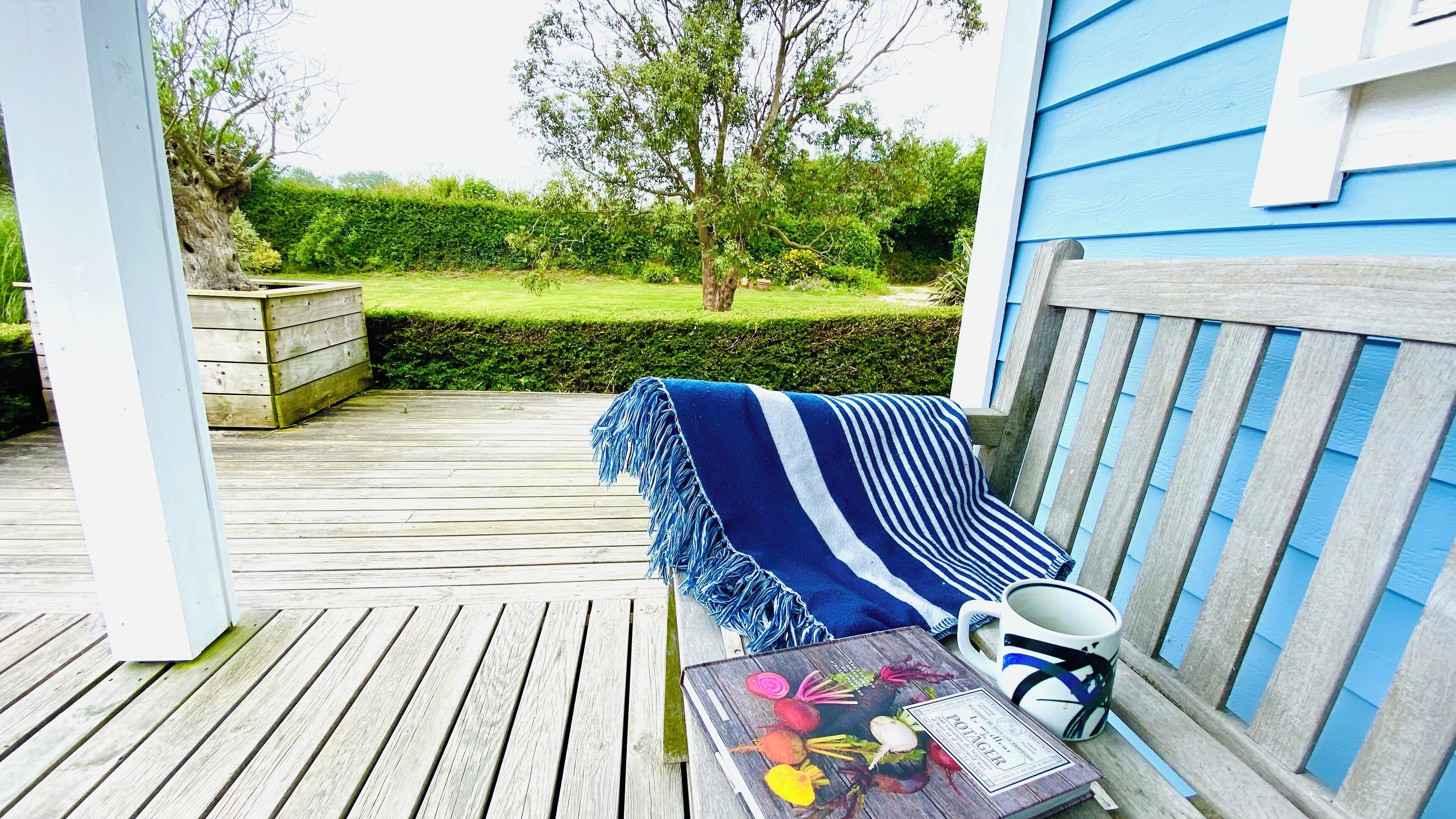 A wooden deck with a blue-striped blanket and a table with a floral book and a mug.