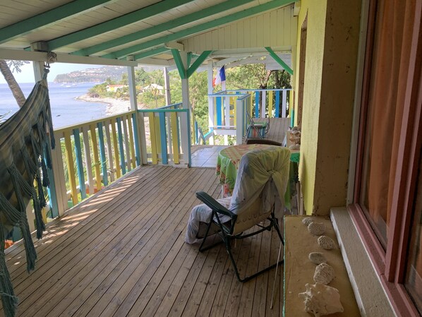 A wooden deck with a hammock, a covered table, and a chair. A view of the sea and buildings is visible through the railing.