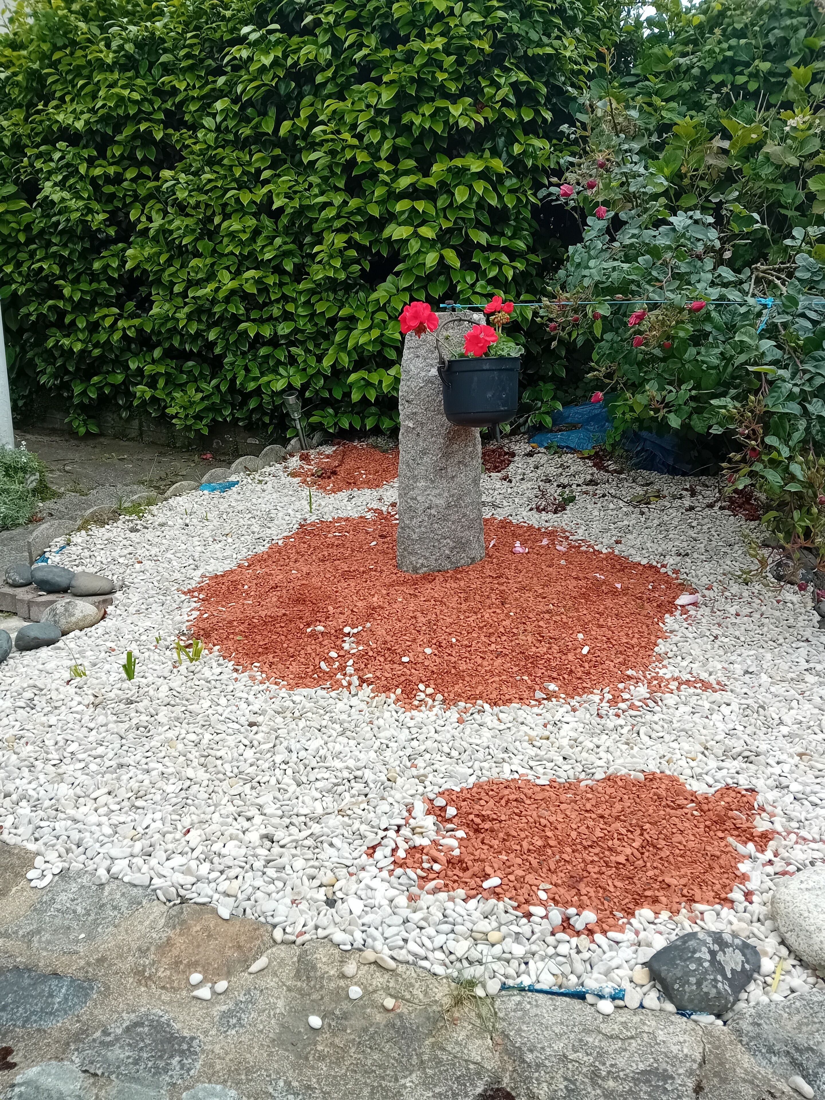 A garden with a central stone pillar, red and white gravel, and green foliage in the background.