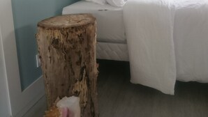 A bedroom with a bed, white pillows, a wooden stool, and a wall with a light switch. - Rental charming T2 Vendée 300m from the beach and 100 m from the forest (Saint-Jean-De-Monts)