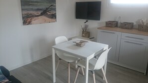 A small, clean room with a white table and chairs, a flat-screen TV mounted on the wall, and a window providing natural light. - Rental charming T2 Vendée 300m from the beach and 100 m from the forest (Saint-Jean-De-Monts)