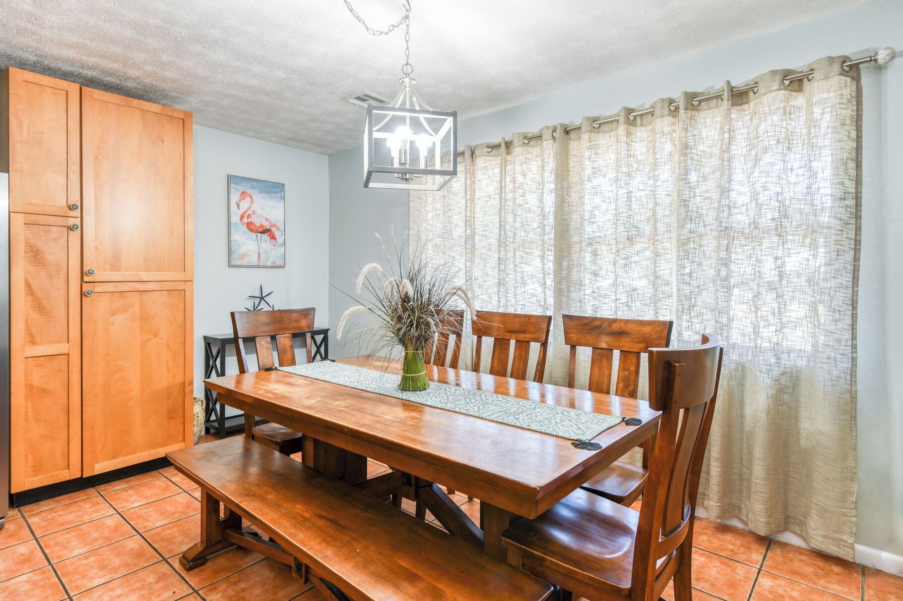 A dining area with a wooden table and chairs, a hanging light fixture, and a window with curtains.