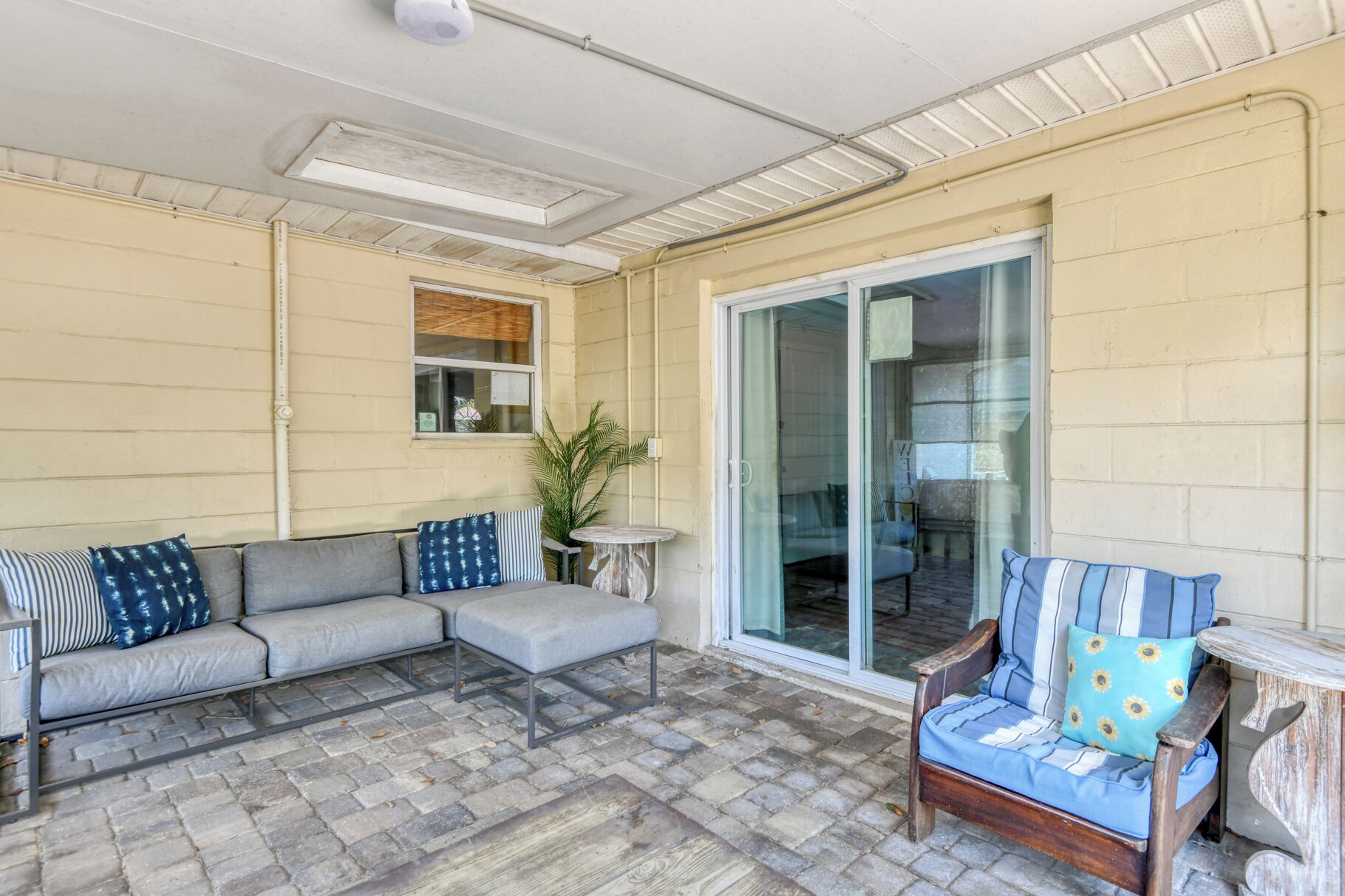 A covered patio area with a grey sofa, a wooden chair, and a small table.
