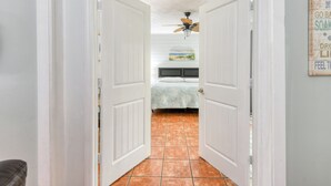 A hallway leading to a bedroom with a bed, a ceiling fan, and a beach-themed painting on the wall.