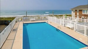 A rectangular pool with a white fence and chairs, overlooking a beach with waves. - Ocean Side Beach Condo in Family Friendly Community!  Oceanfront pool!! (Pine Knoll Shores)
