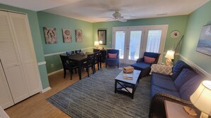A living room with a dining area, a ceiling fan, and artwork on the walls. - Ocean Side Beach Condo in Family Friendly Community!  Oceanfront pool!! (Pine Knoll Shores)